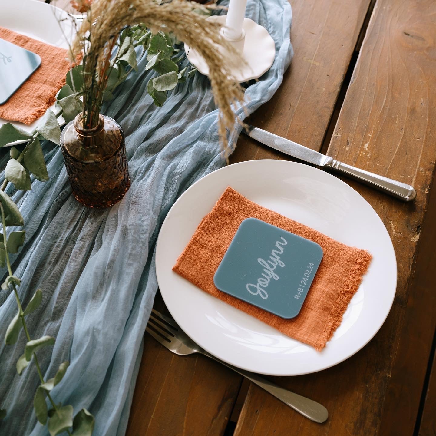 Table setting at a wedding with a plate, orange napkin, and acrylic custom name coaster on a wooden table.