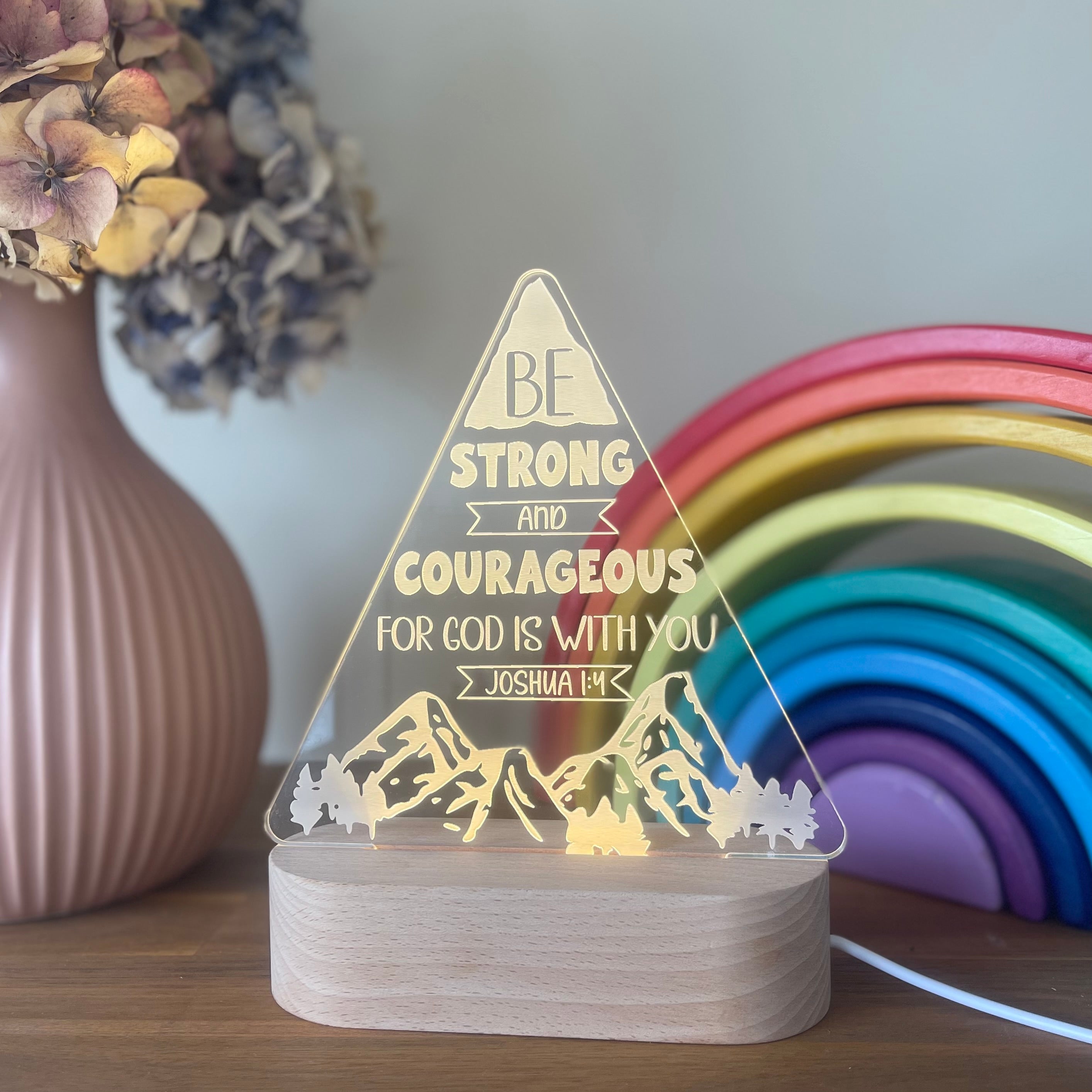 Decorative acrylic night light with bible verse and mountain design, placed on a surface with a rainbow and flowers in the background in a kids room