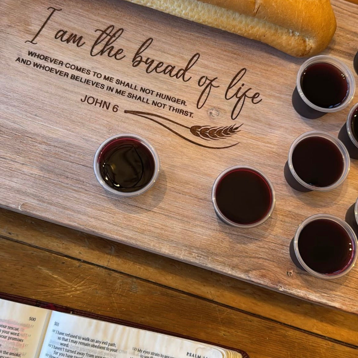 Wooden serving board with bread, wine, and a Bible on a wooden surface with an engraving that reads "I am the bread of life. whoever comes to me shall not hunger, and whoever believes in me shall not thirst"