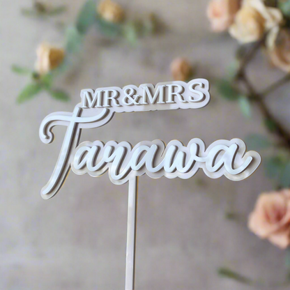 A custom double-layered, acrylic cake topper with 'Mr & Mrs' written in a script font, placed on a wedding cake with a blurred background of flowers.